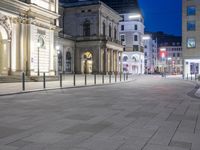 people walking on the pavement of a city street at night time with buildings in the background