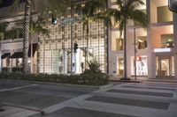 a large glass building with palm trees and buildings at night time in front of it