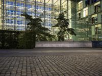 the clock is displayed in front of some tall buildings behind a tree lined street with shrubs