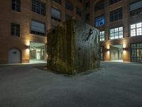 the large sculpture stands outside of a brick building by night in the dark lighted courtyard