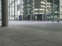 a sidewalk next to a building with tall windows and lights at night, on which are three pillars