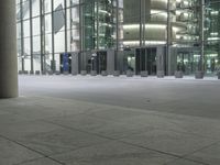 a sidewalk next to a building with tall windows and lights at night, on which are three pillars