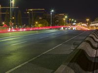 an empty street is seen at night with red light trails in the distance and traffic trails behind