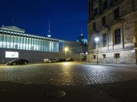 a large stone street with a black car and tall building at night time, behind them is a small streetlight