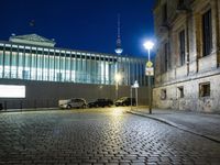 a large stone street with a black car and tall building at night time, behind them is a small streetlight