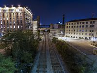 a night time shot of the tracks going past a building complex with other buildings and roads