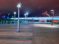 a night scene showing several street lights in a plaza with a bridge and clouds behind