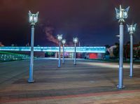 a night scene showing several street lights in a plaza with a bridge and clouds behind