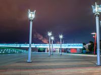 a night scene showing several street lights in a plaza with a bridge and clouds behind