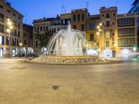 an empty area with many people near a fountain and buildings at dusk in the city
