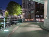 a walkway in front of buildings with a metal railing at night, next to some cars driving on a city street