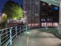 a walkway in front of buildings with a metal railing at night, next to some cars driving on a city street