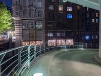 a walkway in front of buildings with a metal railing at night, next to some cars driving on a city street