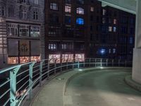 a walkway in front of buildings with a metal railing at night, next to some cars driving on a city street