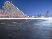 an empty parking lot near a roller coaster at night time, with long exposure of light coming from cars