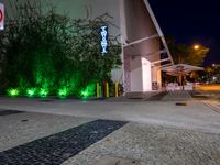 the outside of a building with green lights at night and a sidewalk with plants growing on the sides