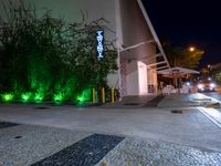 the outside of a building with green lights at night and a sidewalk with plants growing on the sides