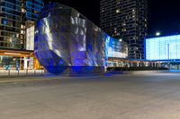 people are walking around by modern architecture at night, some are lights on, in the street