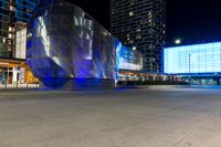 people are walking around by modern architecture at night, some are lights on, in the street