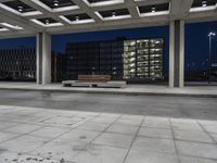 a bench sitting under an overhang at night in a city park area surrounded by concrete columns