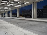 a bench sitting under an overhang at night in a city park area surrounded by concrete columns