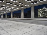 a bench sitting under an overhang at night in a city park area surrounded by concrete columns