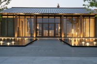 the entrance to a glass house with lights shining on it in front of a concrete walkway