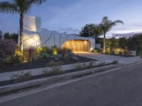 a house at night with lights on the side and palm trees near by in front of the home