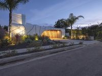 a house at night with lights on the side and palm trees near by in front of the home