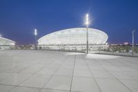 an empty walkway with light poles outside and the stadium building at night in the background