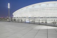 an empty walkway with light poles outside and the stadium building at night in the background