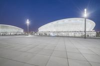 an empty walkway with light poles outside and the stadium building at night in the background