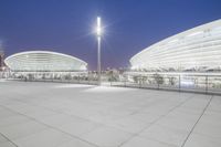 an empty walkway with light poles outside and the stadium building at night in the background