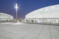 an empty walkway with light poles outside and the stadium building at night in the background