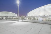 an empty walkway with light poles outside and the stadium building at night in the background