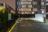 a city building with lights on a rain soaked sidewalk next to a fence and a parking lot