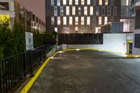 a city building with lights on a rain soaked sidewalk next to a fence and a parking lot