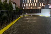 a city building with lights on a rain soaked sidewalk next to a fence and a parking lot