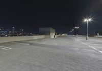 an empty parking lot with several lights on top of it at night with buildings in the background