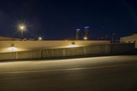 an empty road near two tall buildings with street lamps in the middle of it at night