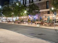 an outdoor space is lit up at night, and there are tables and benches along the sidewalk