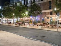 an outdoor space is lit up at night, and there are tables and benches along the sidewalk