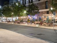 an outdoor space is lit up at night, and there are tables and benches along the sidewalk