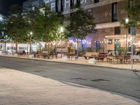 an outdoor space is lit up at night, and there are tables and benches along the sidewalk