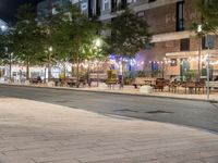 an outdoor space is lit up at night, and there are tables and benches along the sidewalk