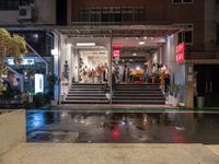 a small courtyard with stairs and the entrance of a store on a wet street with a few people walking and sitting