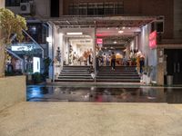 a small courtyard with stairs and the entrance of a store on a wet street with a few people walking and sitting