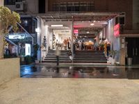 a small courtyard with stairs and the entrance of a store on a wet street with a few people walking and sitting