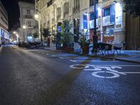 an empty city street lined with buildings at night time as people are seated at tables and talking on the pavement