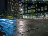 a wet street that has some buildings and cars in it at night time with water puddles on the street